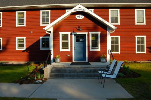 A red wooden building with a welcoming entrance, featuring steps, a blue door, and two white chairs outside.