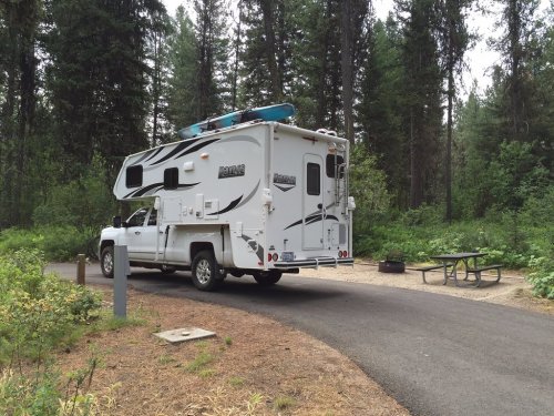 A white camper truck parked at a campsite surrounded by tall trees and greenery. A picnic table is nearby.