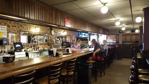 A cozy bar interior with wooden accents, patrons sitting at the counter, and a well-stocked bar in the background.