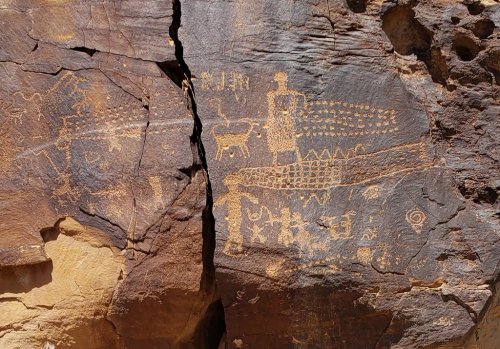Petroglyphs on a rock surface depicting human figures, animals, and geometric patterns in earthy tones.