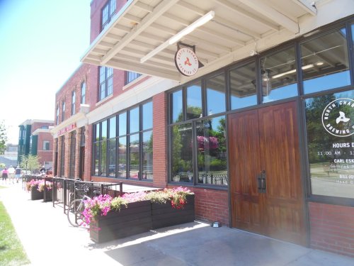 Exterior of a brick building with large windows, featuring a sign and flower planters along the sidewalk.