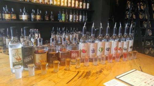 A wooden table displays various bottles of spirits and shot glasses in a tasting room setting.
