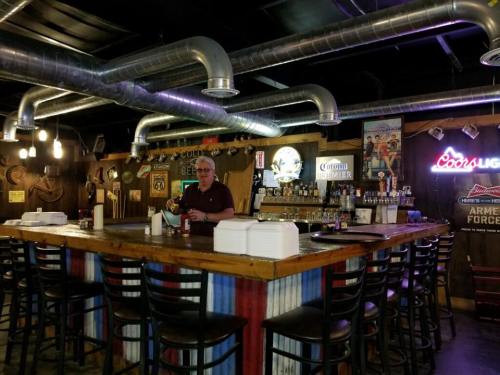 A bartender stands at a rustic bar with metal accents and various liquor bottles in a cozy, dimly lit setting.