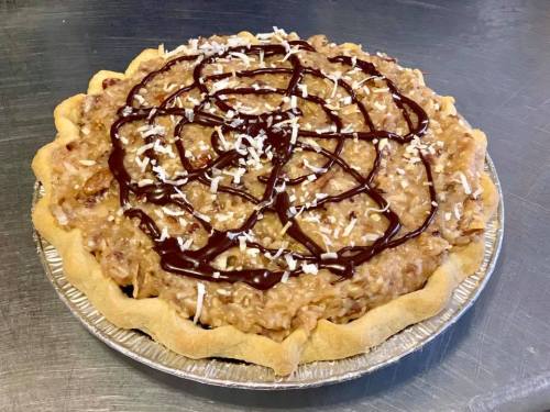 A delicious pie topped with coconut, chocolate drizzle, and a golden crust, sitting on a silver pie plate.