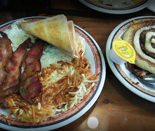 A plate of breakfast featuring bacon, hash browns, eggs, toast, and a cinnamon roll with icing on the side.