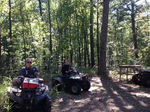 Two people on ATVs in a wooded area, surrounded by trees and greenery, with a wooden table in the background.