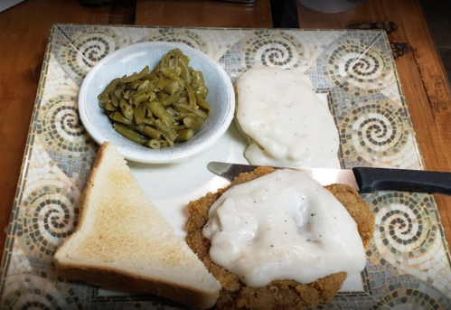 A plate with fried chicken topped with gravy, green beans, a slice of bread, and two biscuits.