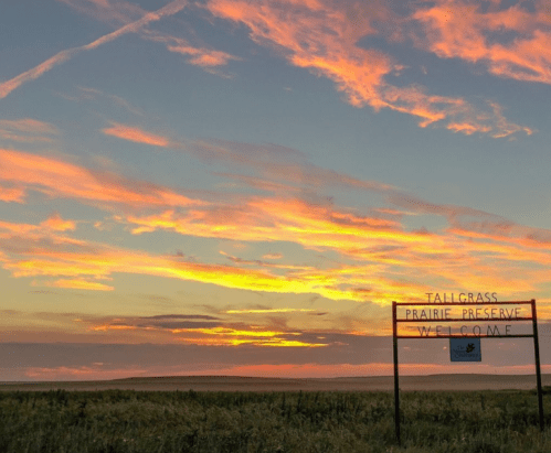 A vibrant sunset over a grassy landscape with a sign reading "Welcome to Tallgrass Prairie Preserve."