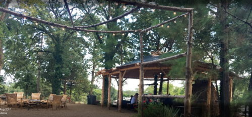A rustic outdoor pavilion surrounded by trees, with string lights and wooden seating.