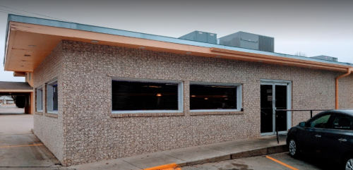 A single-story building with a textured exterior, large windows, and a door, parked car in front.