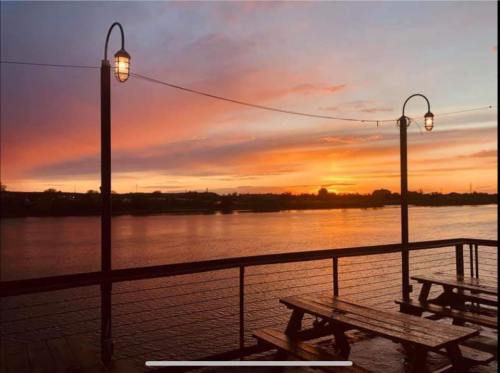 Sunset over a calm river, with picnic tables and lights on a wooden deck, creating a serene atmosphere.