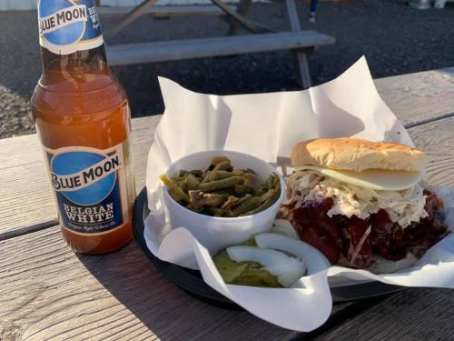 A bottle of Blue Moon beer next to a plate with a pulled pork sandwich, coleslaw, and a side of green beans.