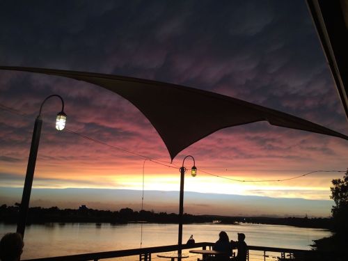 A vibrant sunset over a calm river, with dramatic clouds and silhouettes of people under a shaded patio.