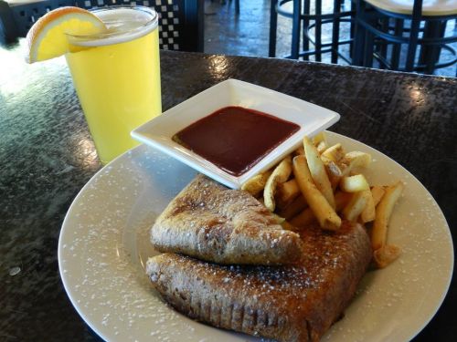 A plate with two stuffed French toast slices, fries, and a small bowl of sauce, alongside a glass of yellow drink.
