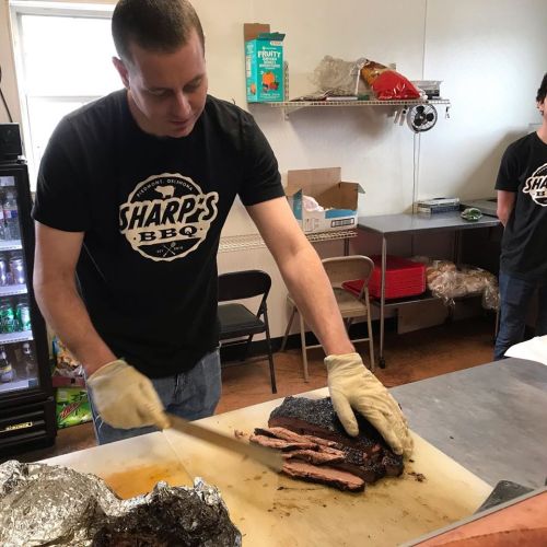 A person in a BBQ shirt slices brisket on a cutting board in a kitchen setting.