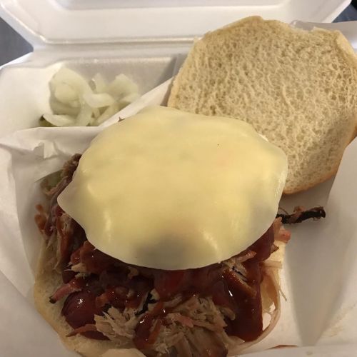 A pulled pork sandwich topped with melted cheese and barbecue sauce, served with pickles and onions on the side.