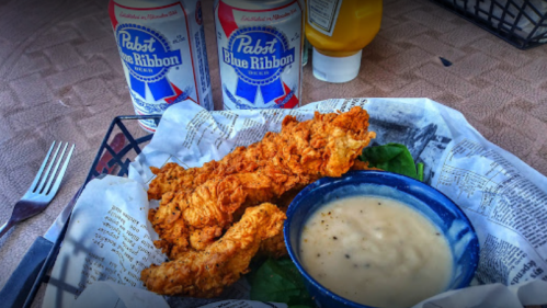 A basket of fried chicken strips with a bowl of gravy, accompanied by two cans of Pabst Blue Ribbon beer.