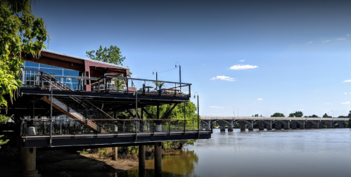 A riverside restaurant with a deck, overlooking a calm river and a bridge under a clear blue sky.