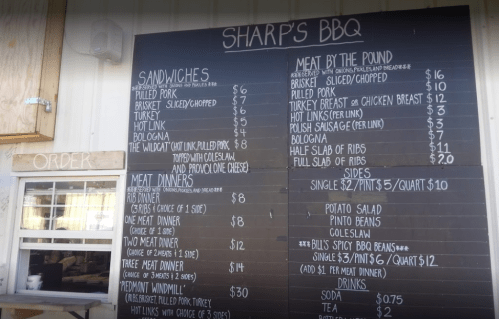 Menu board at Sharp's BBQ listing sandwiches, meat by the pound, and meat dinners with prices.