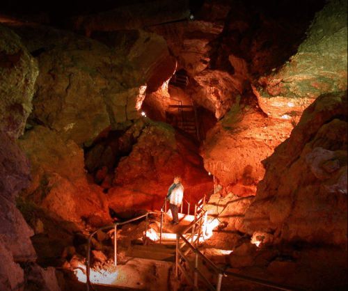 A person stands on a staircase inside a dimly lit cave, surrounded by colorful rock formations and illuminated pathways.