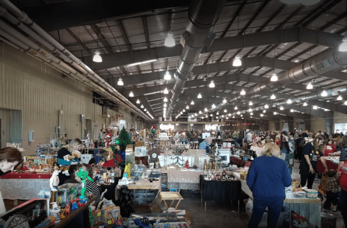 A bustling indoor market with various vendor tables, shoppers browsing, and colorful items on display.