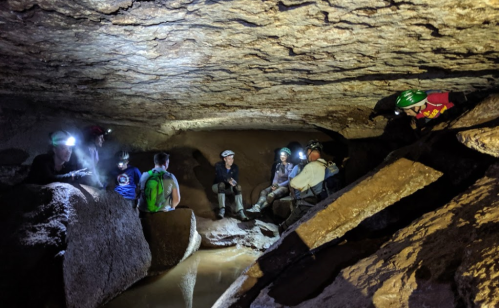 A group of cavers with headlamps sits in a rocky cave, surrounded by boulders and a small pool of water.