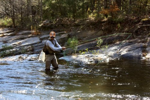 A person fishing in a river, wearing waders and a hat, with trees and rocks in the background.