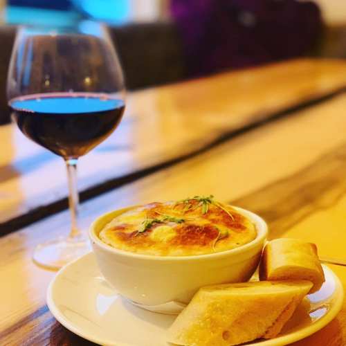 A bowl of baked dish topped with cheese, served with bread, alongside a glass of red wine on a wooden table.