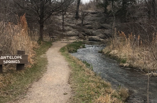 A winding dirt path beside a stream, with a sign for Antelope Springs and sparse trees in the background.