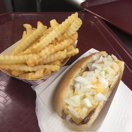 A hot dog topped with onions and cheese, served with a side of crinkle-cut fries on a red tray.