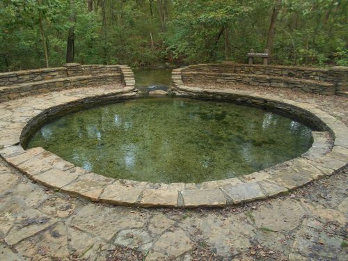A circular stone pool surrounded by trees, reflecting the greenery above in calm water.