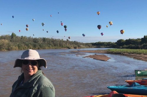 A woman in a hat smiles by a river, with colorful hot air balloons floating in the sky above.