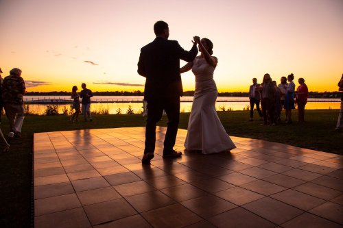 A couple dances on a wooden floor at sunset, surrounded by guests enjoying the evening by the water.