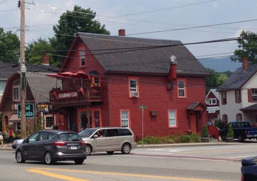 A red two-story building with a cafe sign, surrounded by trees and parked cars on a sunny day.