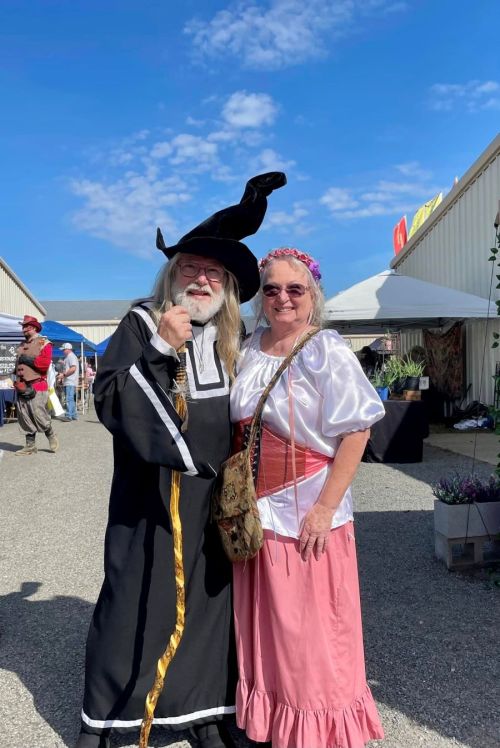 A man in a wizard costume and a woman in a historical dress pose together at an outdoor event under a blue sky.