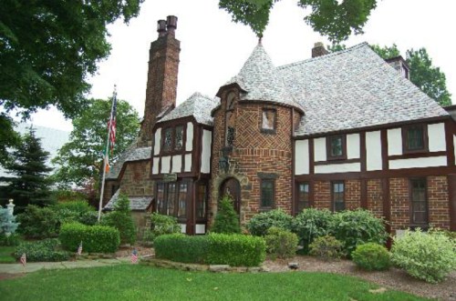 A charming brick and timber house with a steep roof, surrounded by lush greenery and a flagpole in the front yard.