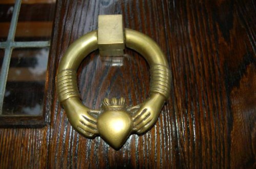 A decorative brass door knocker shaped like two hands holding a heart, mounted on a wooden door.