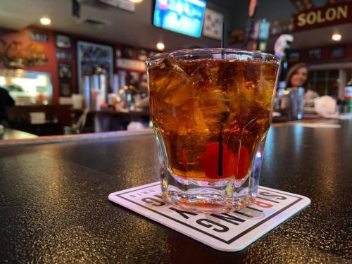 A close-up of a glass of whiskey with ice and a cherry, sitting on a coaster at a bar. Background shows a dimly lit bar scene.