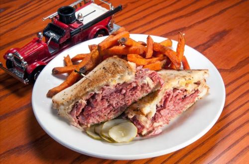 A plate with a sandwich filled with meat, pickles, and sweet potato fries, alongside a toy fire truck.