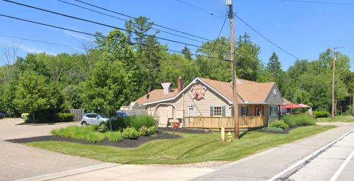 A small, light-colored building with a porch, surrounded by greenery, on a sunny day with clear blue skies.