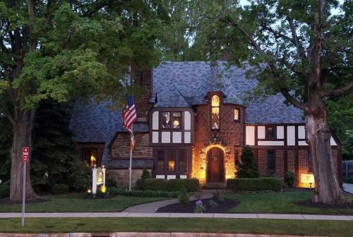 A charming two-story house with a mix of brick and wood, illuminated at dusk, featuring an American flag and landscaped yard.