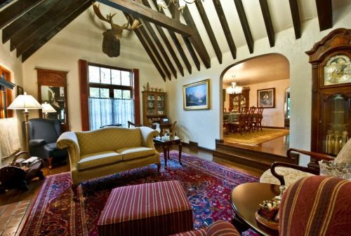 Cozy living room with high wooden beams, a mounted deer head, elegant furniture, and a view into a dining area.