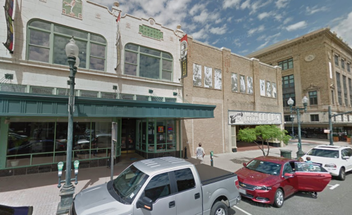 Historic building facade with banners, parked cars, and a person walking on a sunny day in a downtown area.