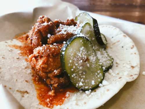 A tortilla topped with crispy fried chicken, sliced pickles, and crumbled cheese, drizzled with sauce.