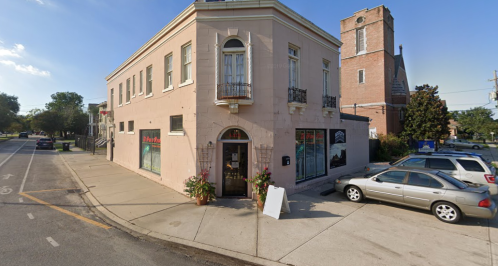 A pink building with balconies, featuring storefronts and parked cars on a sunny street corner.