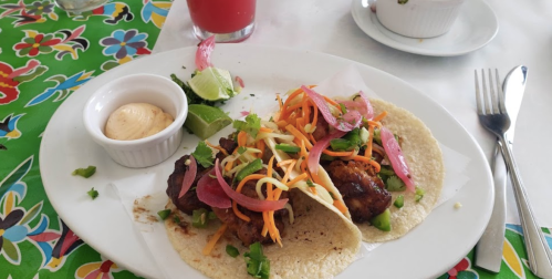 Two tacos filled with grilled meat, topped with colorful vegetables, served with lime and a side of sauce.