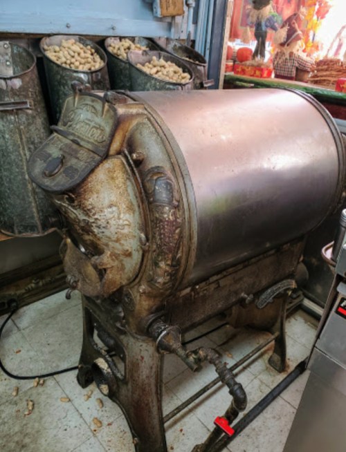 An antique peanut roaster with a metallic body, featuring a vintage design and a worn cap, displayed in a shop.