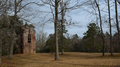 A weathered brick tower stands among bare trees on a grassy landscape under a cloudy sky.