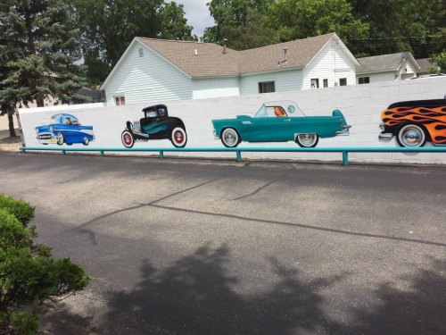 A mural featuring vintage cars painted on a wall beside a parking lot, with a house and trees in the background.