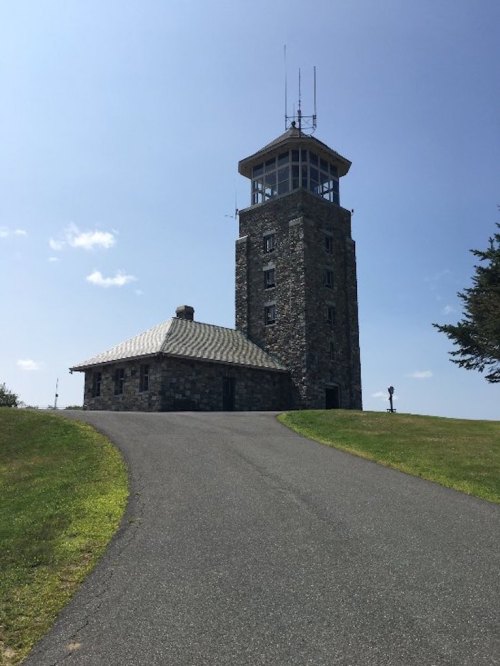 A stone tower with a glass observation deck stands beside a smaller building, set against a clear blue sky.